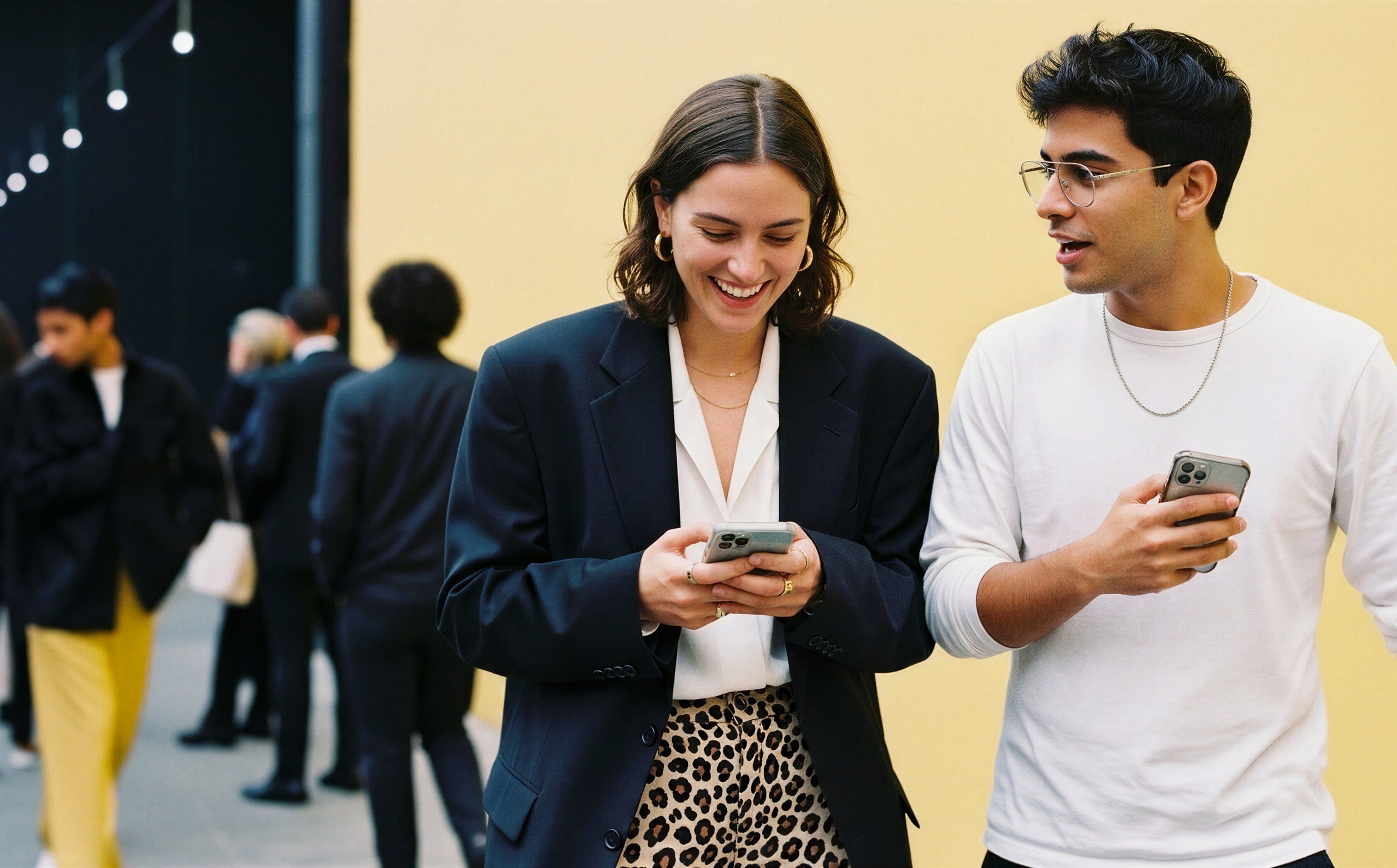 A smiling woman in a maroon blazer and a man in a black cardigan standing close together, looking down at a smartphone screen